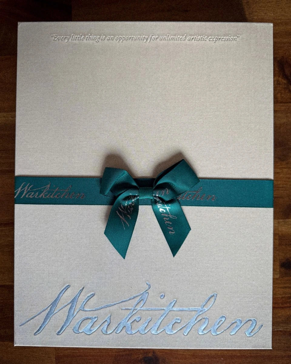 Linen box with midnight green grosgrain ribbon with "Every little thing is an opportunity for unlimited artistic expression" debossed on the lid